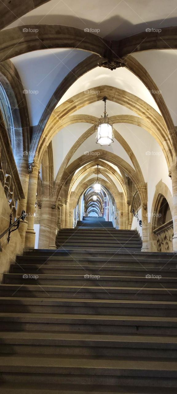 This lavish hallway with stairs inside the „Alter Hof“ in „Munich“ has a cathedral ceiling intersected by arched windows below. Arches also extend upwards diagonally and meet at a top center point in each section. 2023. Hypnotic Productions