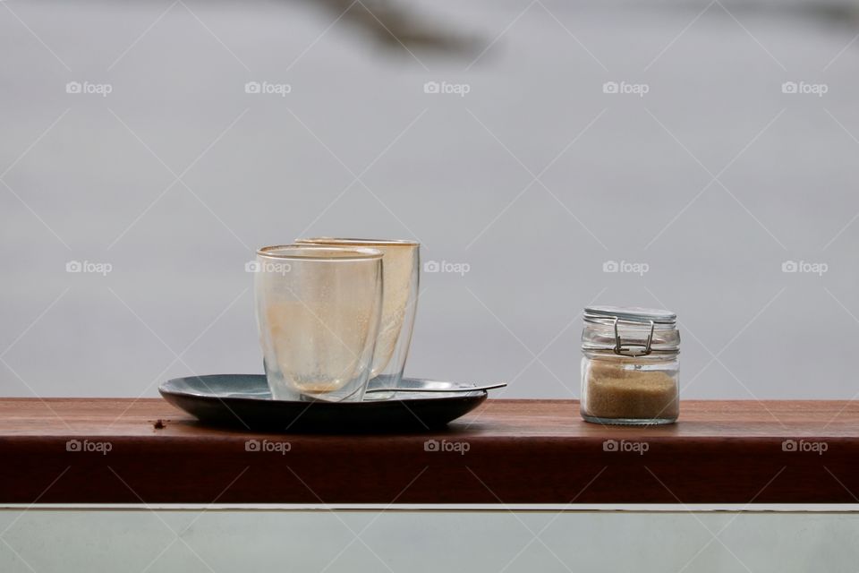 Two empty iced cappuccino glasses on black plate on ledge with mason jar of sugar on outdoor restaurant patio overlooking ocean, early morning, still life with copy text space