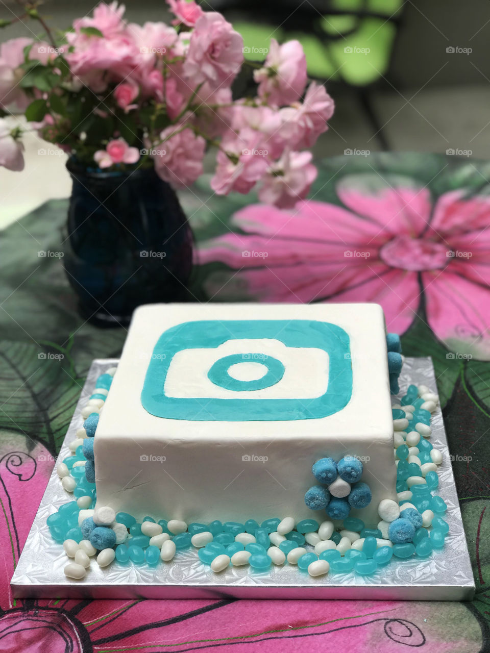 A foap wedding cake with the logo made of white & turquoise fondant. Daisies of turquoise gummies & marshmallows on the side of the cake & turquoise & white jelly beans on the cake base.  All on a green & pink table cloth with a vase of pink roses. 