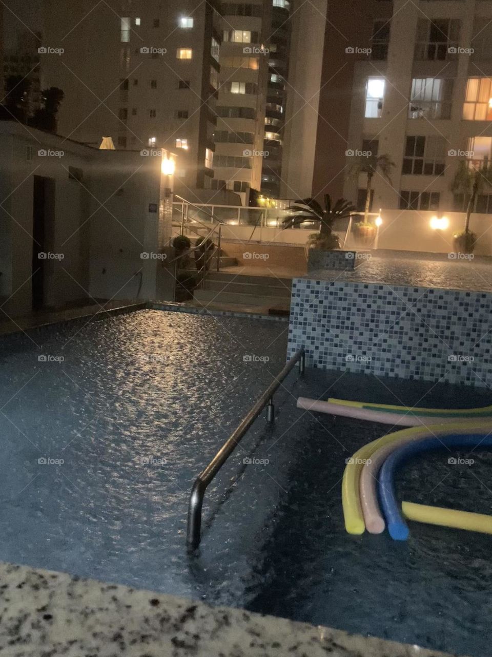 Prédio com piscina. Do outro lado da rua muro com luzes. Luzes no interior dos apartamentos vistas pelas janelas.