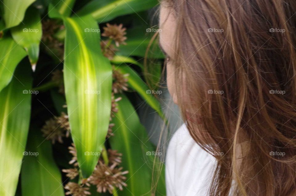 Young woman in a white blouse and face hidden in her brown hair next to a tropical plant.