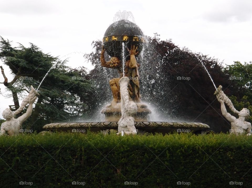 A decorative fountain with a gold statue holding a globe that has astrology symbols on it on his shoulders and three white statues blowing water from cornucopias onto him on a summer day in England.