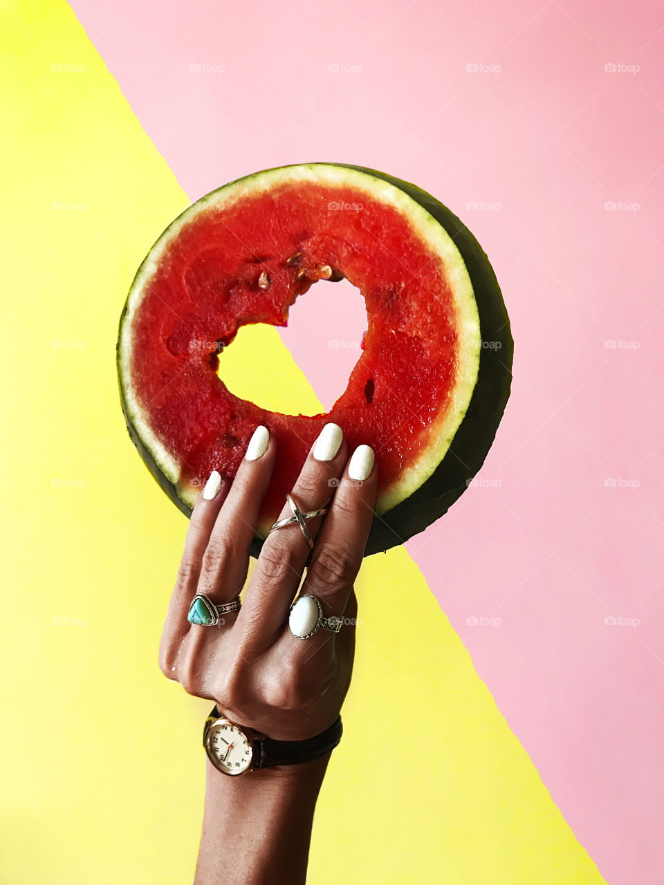 Female hand with white nails and fashionable accessories holding a red watermelon with a heart shaped hole on colorful pastel background 