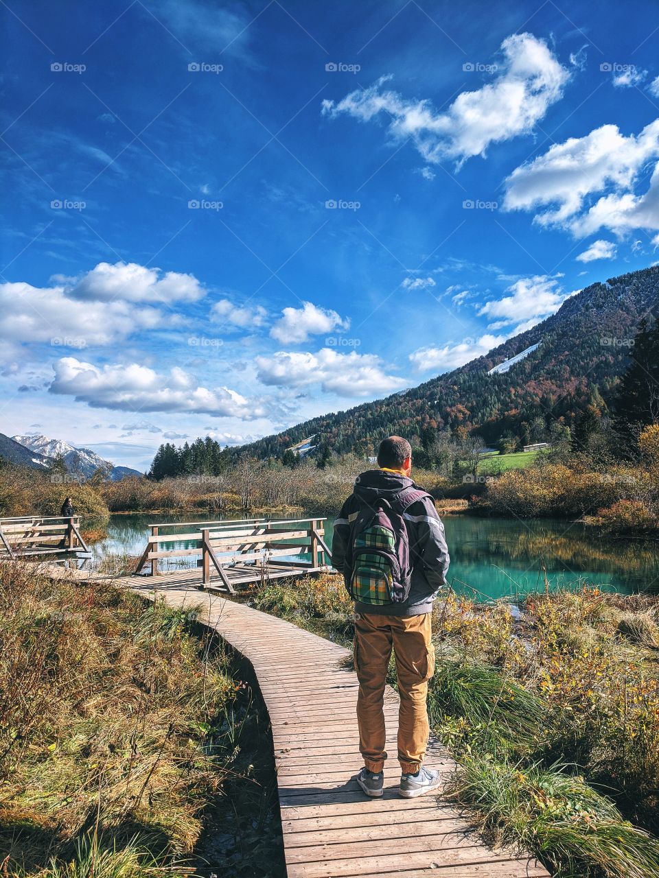 A male traveler stands on the road and admires the view of the mountain peaks above lake.