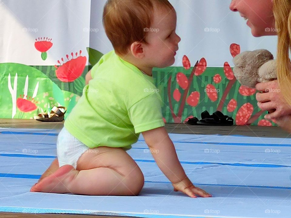 Love: parents and children.  A little boy in a diaper is sitting on the podium.  The boy looks at his smiling mother, who is holding a teddy bear. The connection between baby and mother