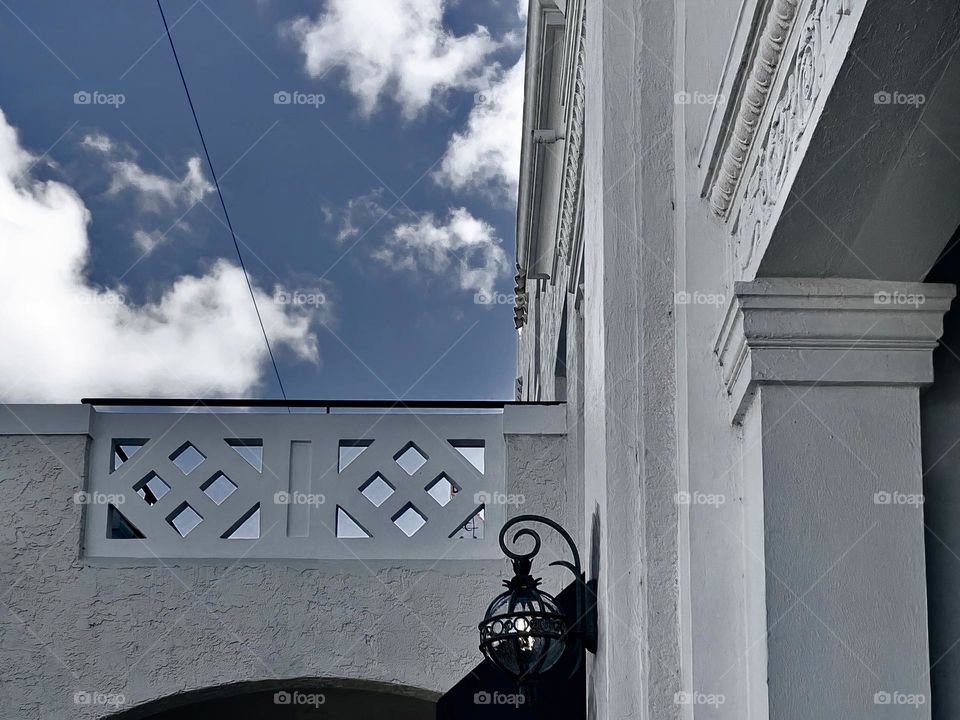 Beautiful details and art carving on white building with architectural built with historical appearance close-up looking upward from the arched doorway and lamp style light fixture to the sky with clouds.