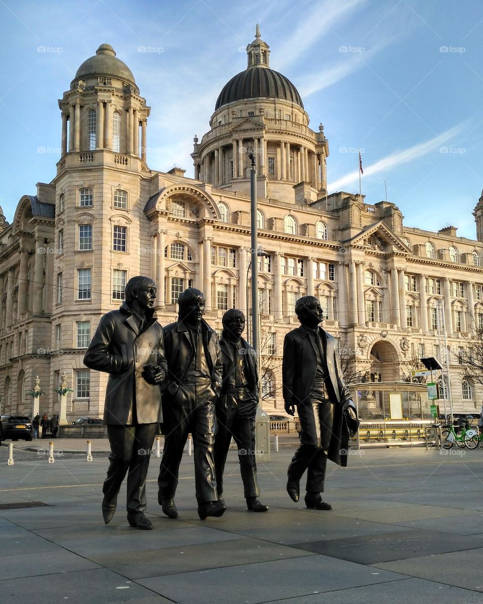 Statue of The Beatles in Liverpool, England.  In the background, the sun shines on one of the emblematic buildings of the city.