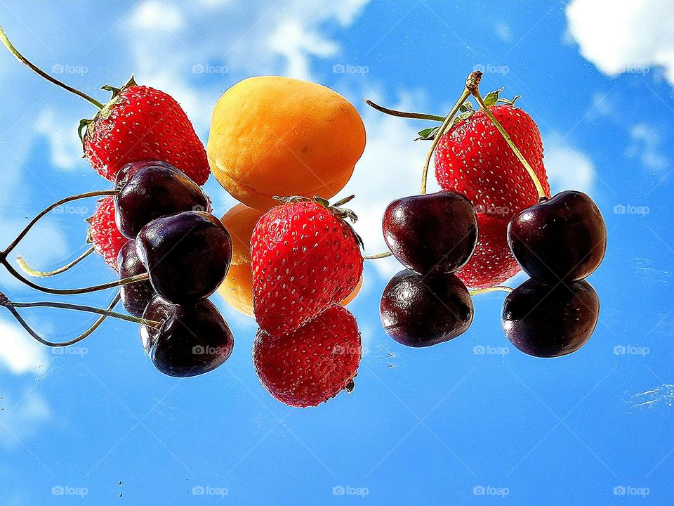 Summer. Time for fruits and berries. On the mirror surface lie: yellow apricot, red strawberries and dark burgundy cherries. Blue sky reflection with white clouds