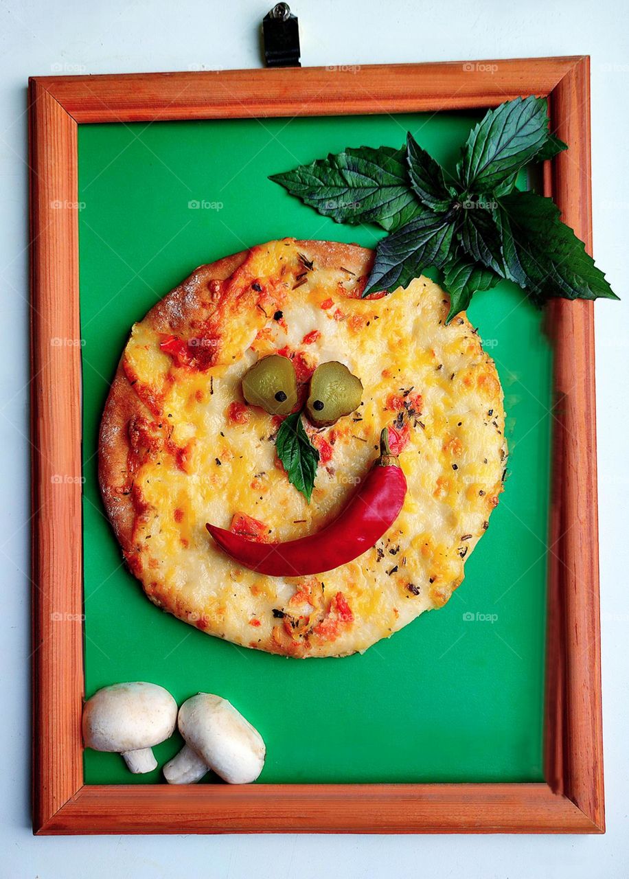 Green color.A wooden frame hangs on a white wall.Framed composition.On a green background a mini pizza with two green circles of pickles in the shape of eyes,with a green leaf of basil-nose and a red chili pepper-smile. Green basil in the top corner.