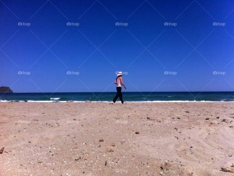 Women walking on beach, wearing a large hat, pink Tshirt and black trousers. 
Deep blue sky, sand with visible small white shells and small waves.