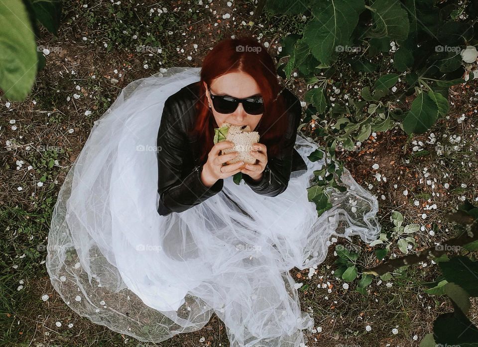 an original portrait of a girl in a black leather jacket, a white fluffy skirt and glasses sitting under a tree and eating a sandwich appetizingly