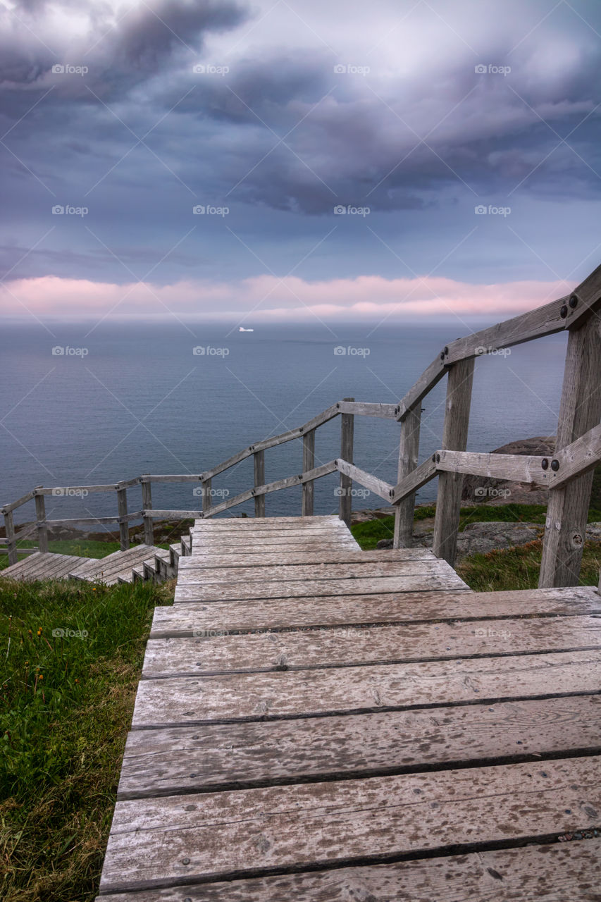 Iceberg floating in the sea under dramatic storm clouds, with a winding wooden staircase leading down to the shore. Signal Hill - St Johns, Newfoundland