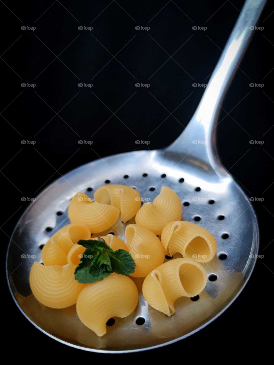 Favorite food. Pasta. Yellow "pipe rigate" pasta rests on a gray metal slotted spoon. Green mint leaves lie on top of the pasta. Black background