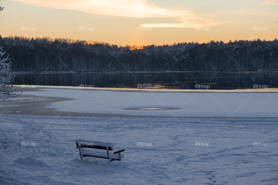 Snowy landscape at sunset, park bench with a nice view of forest and frozen lake covered with snow 