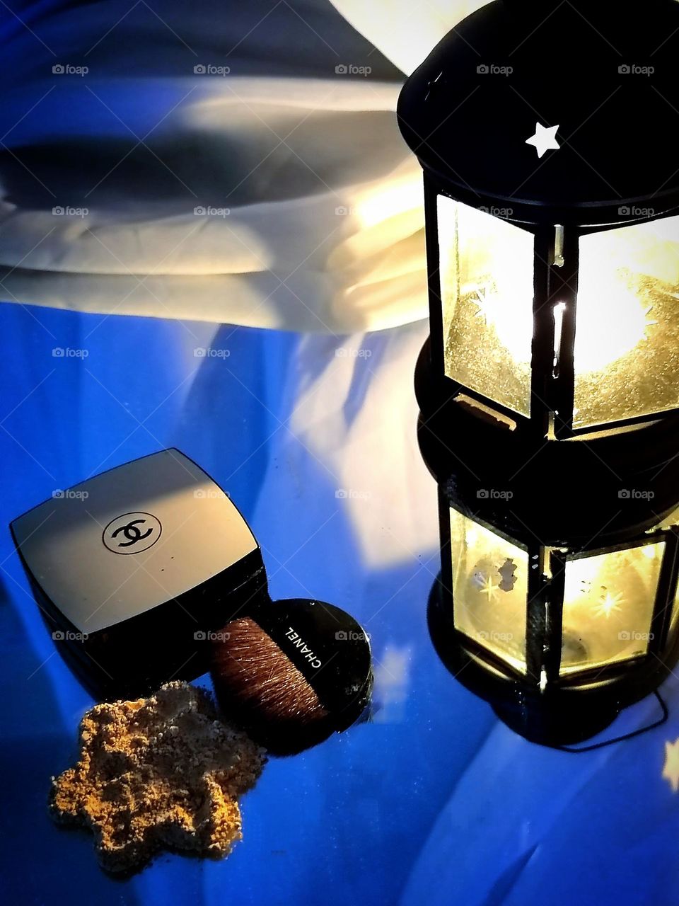 A lantern is lit against a mirror background, the light from which falls on a box with the Chanel logo, on powder and a brush with the inscription "CHANEL". In the background is a white air curtain. Powder Chanel Les Beiges