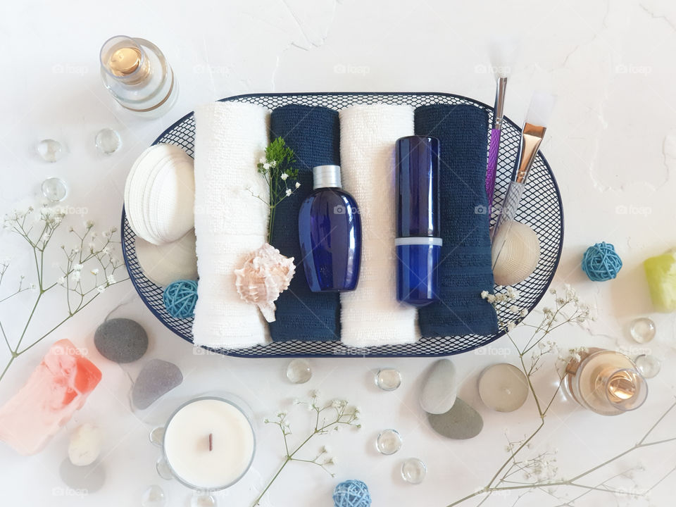 Blue cosmetic bottles on folded towels in a basket with seashells on a white background. Beauty salon, table with creams, soap, towels and candles, top view