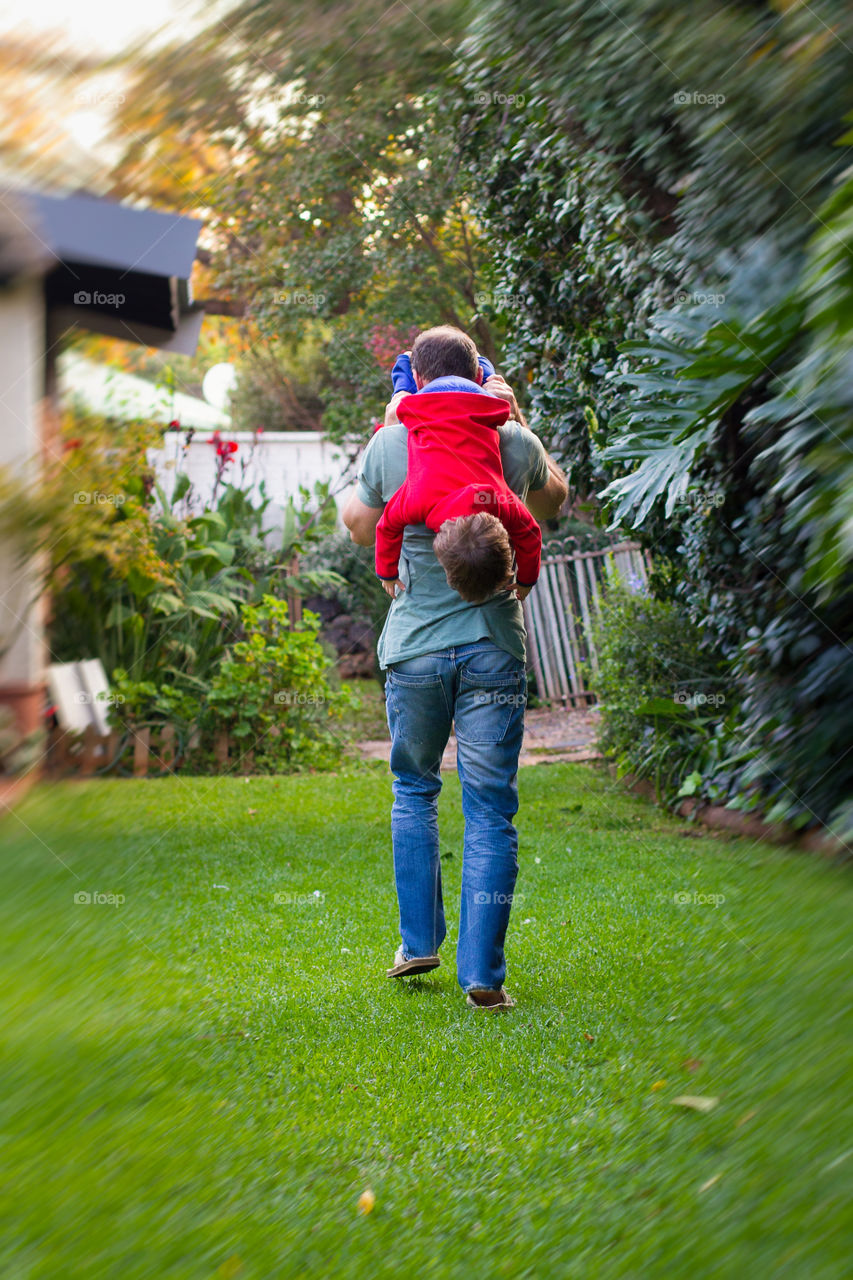 Father and son durong covid lockdown - man walking for exercise around the garden with boy playing and hanging from his shoulders, late afternoon.