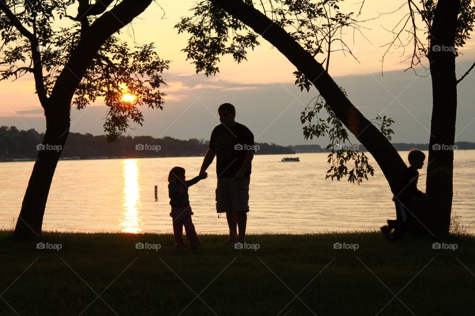 Gorgeous golden bright yellow sun is setting off in the distant horizon with beautiful silhouette of father and daughter holding hands under trees! 