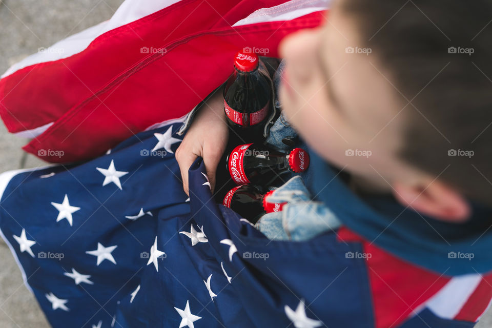 a teenage guy sits on a bench and holds Coca-Cola bottles in his hands, wrapped in an American flag