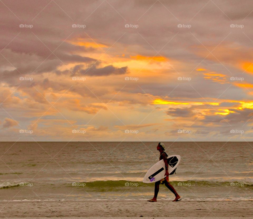 Summer sunset on the beach with a surfer walking by carrying his surfboard 