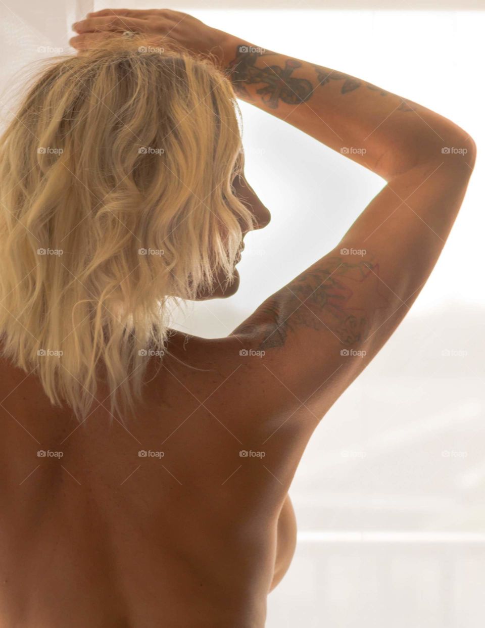 image of nude woman from the back showing her right shoulder and arm raised to her blonde hair which covers her face that is turned to the side all in front of a soft white window curtain
