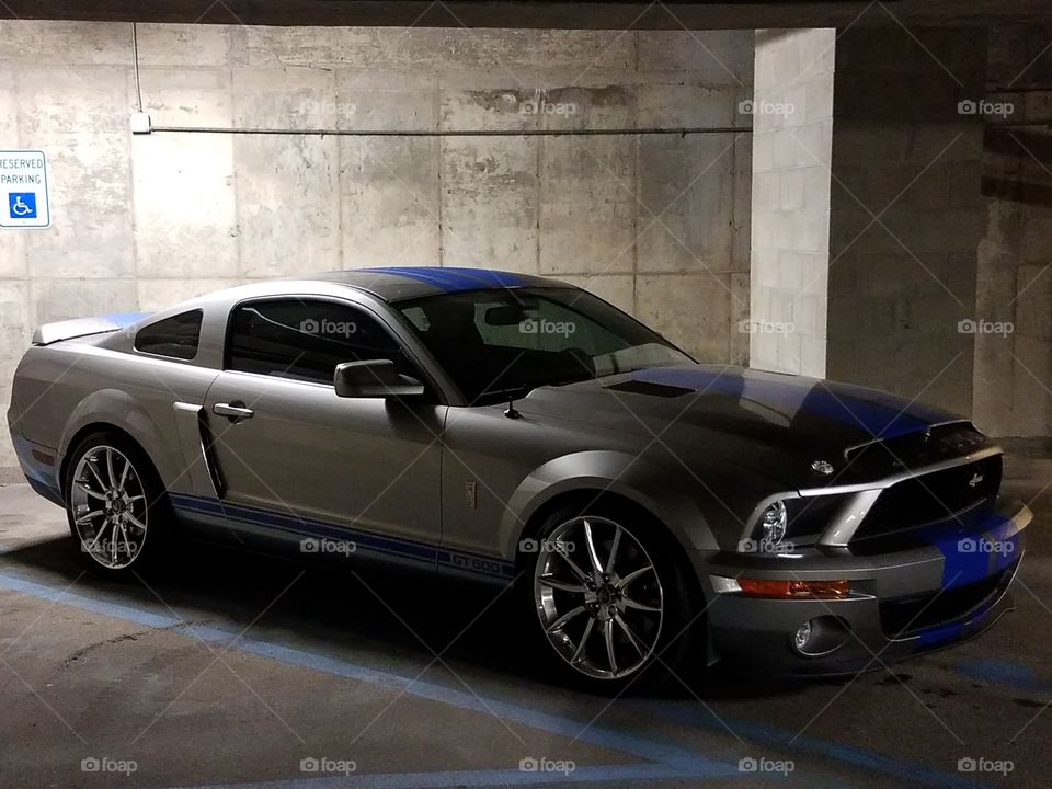 A silver Shelby mustang with blue racing stripes takes up a handicap spot in a garage.