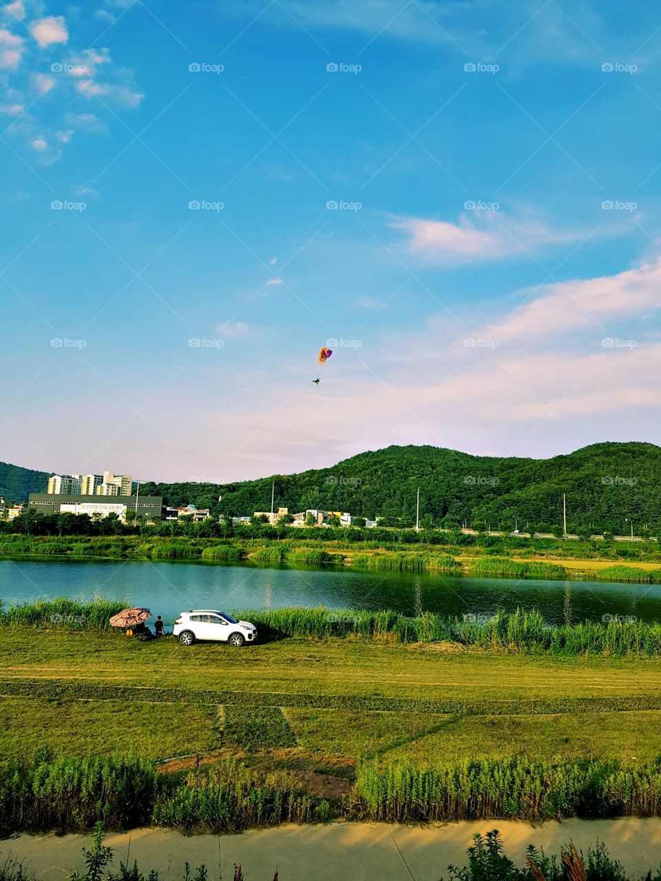 It's a fields where there is a big lake at the front,  buildings and homes.
there is a guy below the car watching the water and the person cross to him who is flying in the sky with a parachute.