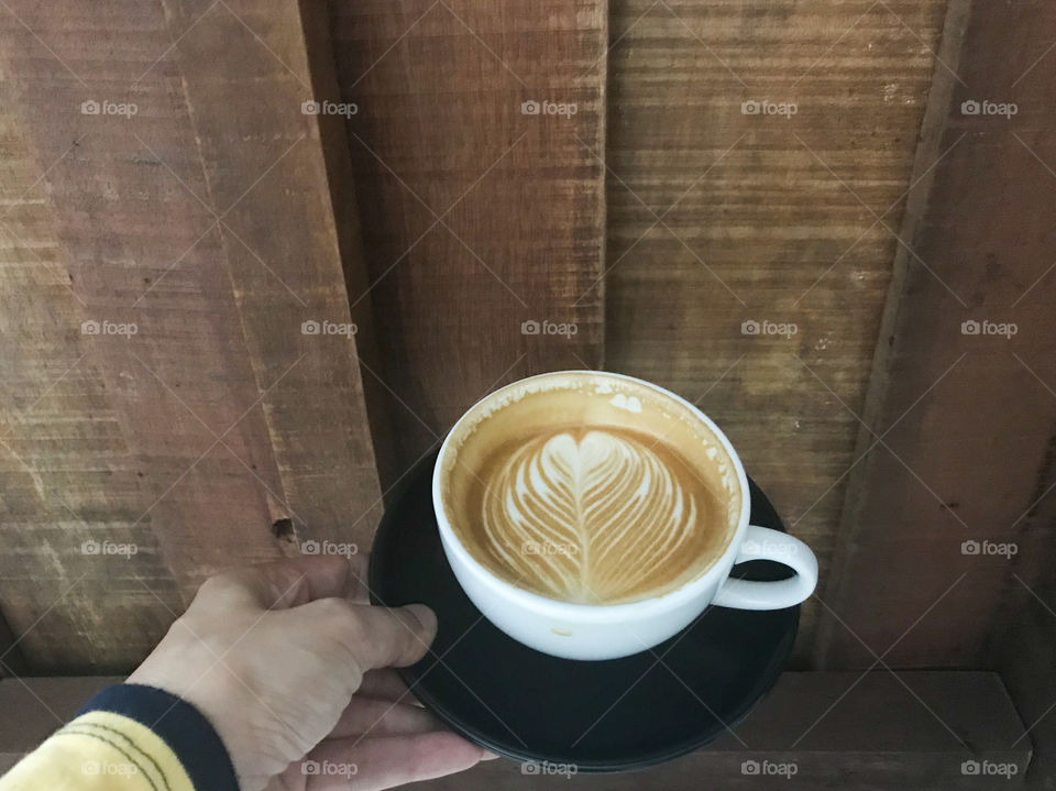 woman wears yellow and dark blue blouse is holding a white cup of hot latte coffee with beautiful flower latte art and brown natural wooden board on background
