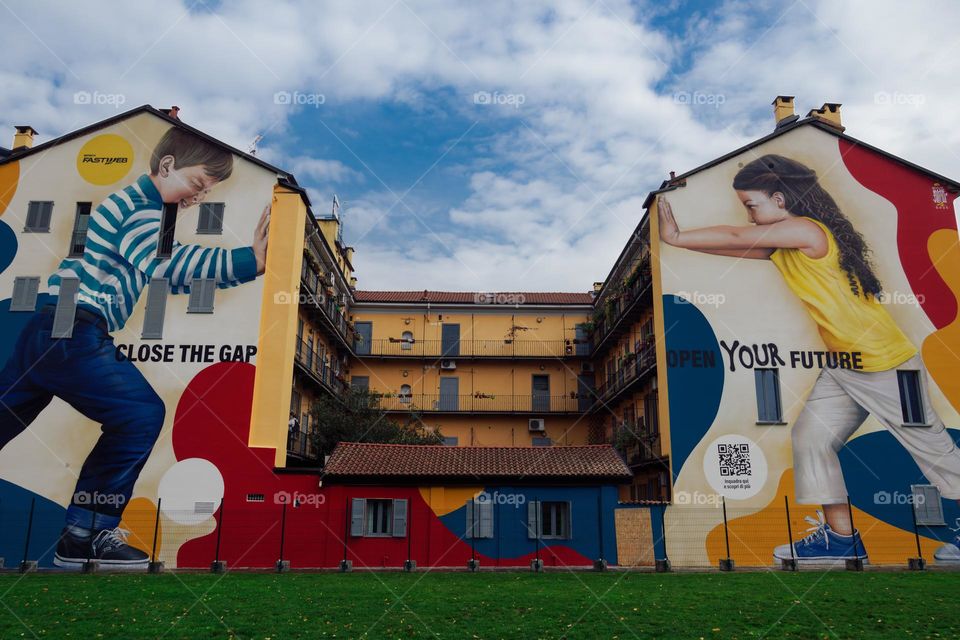 Milan, Italy colorful wall graffiti under blue sky. Vibrant street art mural by  artist Rosk depicting two children pushing a wall with the message Close The Gap, Open Your Future.