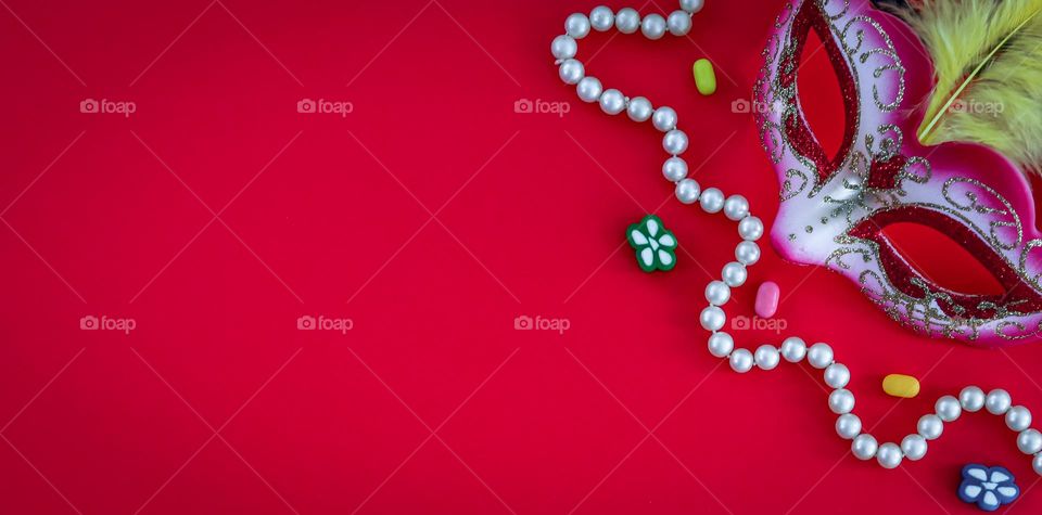 One masquerade mask, a white pearl necklace and colored jelly beans lie on the right on a red background with copy space on the left, flat close-up. Concept for masquerade, festival, carnival, party banner.