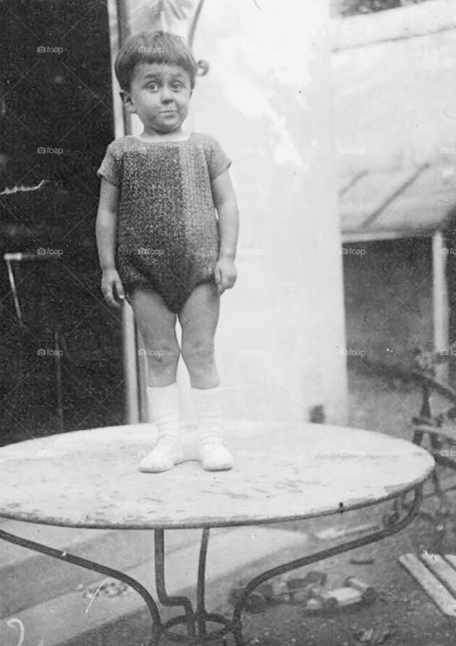 Vintage black and white shot taken in 1924 of a toddler standing on a marble garden table not too impressed by the wool knit bodice knitted by his mother