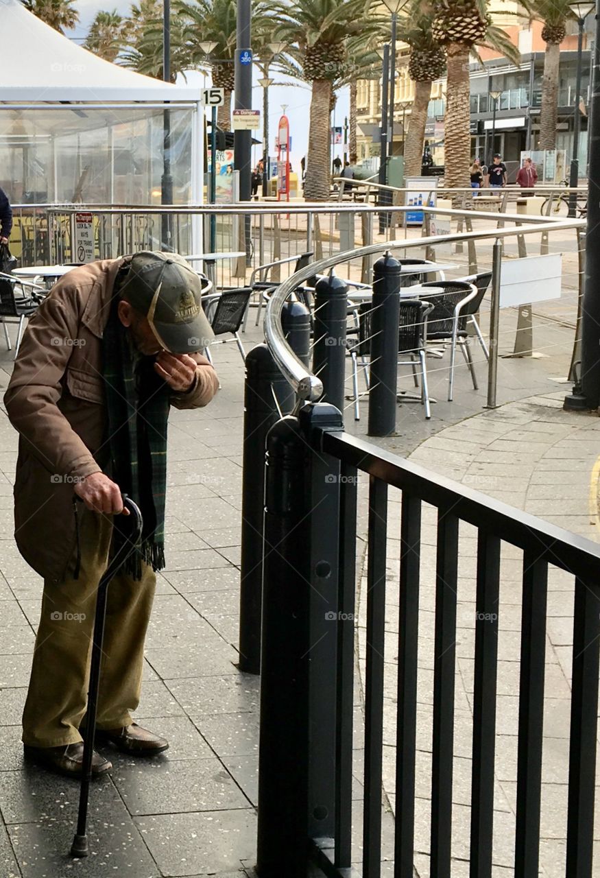 Elderly hunched over man wearing coat with cane and baseball cap in the city 