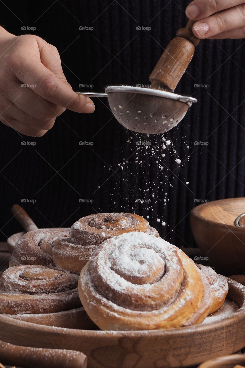 women's hands pour powdered sugar through a sieve on cinnamon rolls