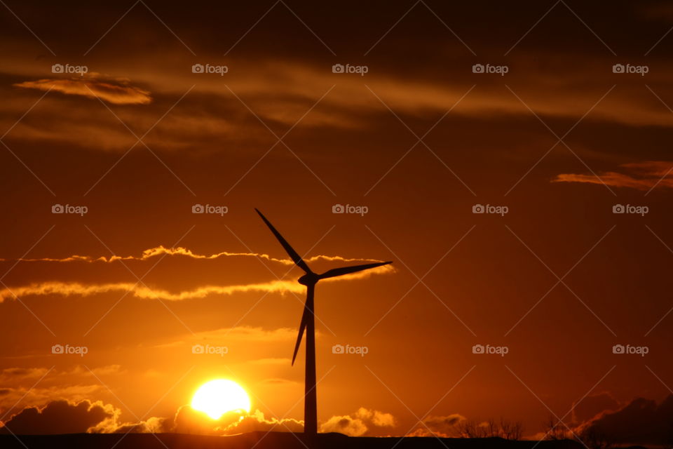 Wind turbine on the horizon at sunset. Orange sky. Bright sun behind a cloud. Picture taken in Salento, Puglia, Italy.