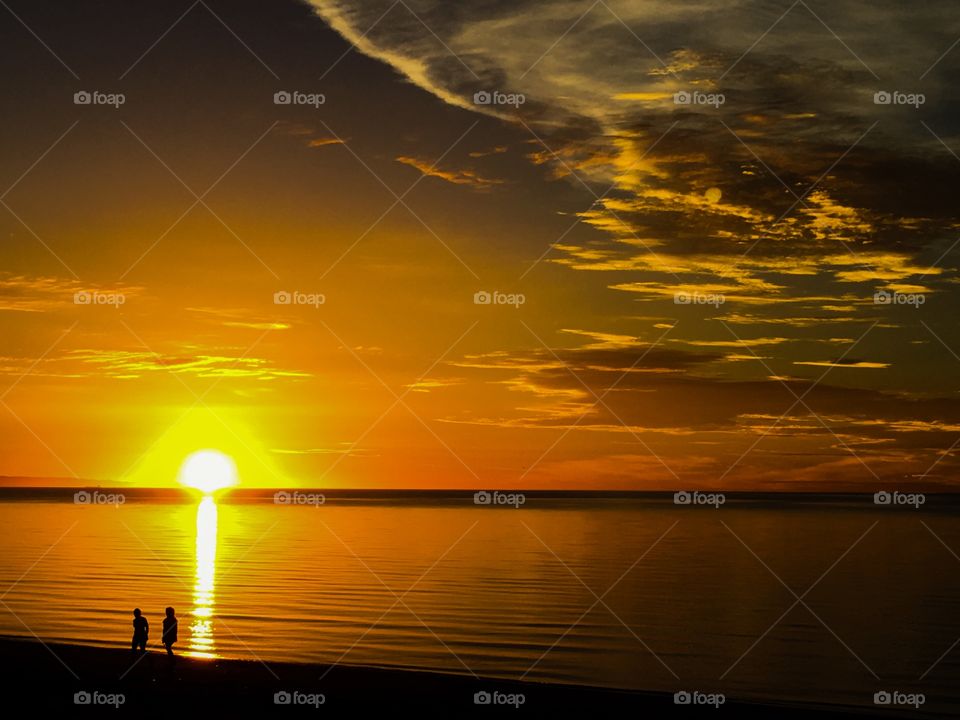 Sun rising on the horizon in a brilliant orange sunrise, sun reflection on calm ocean water and silhouette of couple walking