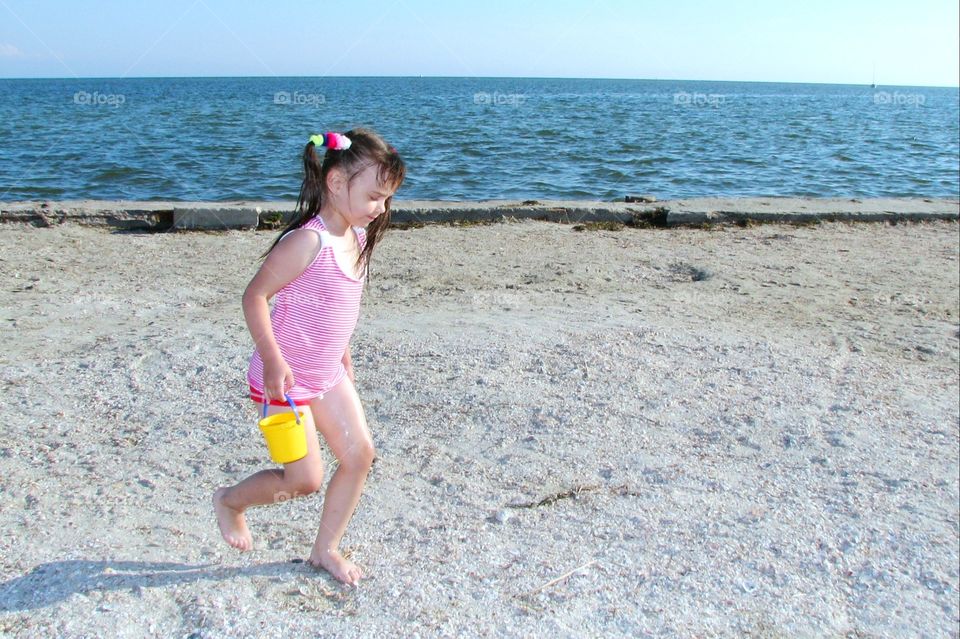 little girl walks along the seashore with a yellow bucket