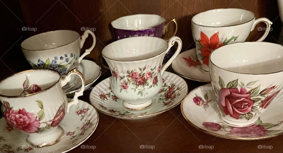 The shelf in the curio cabinet showcases six unique antique tea cups, each with its own distinct design, paired with matching plates. Each cup has a different handle style adding to their individuality. The matching plates are similarly varied.