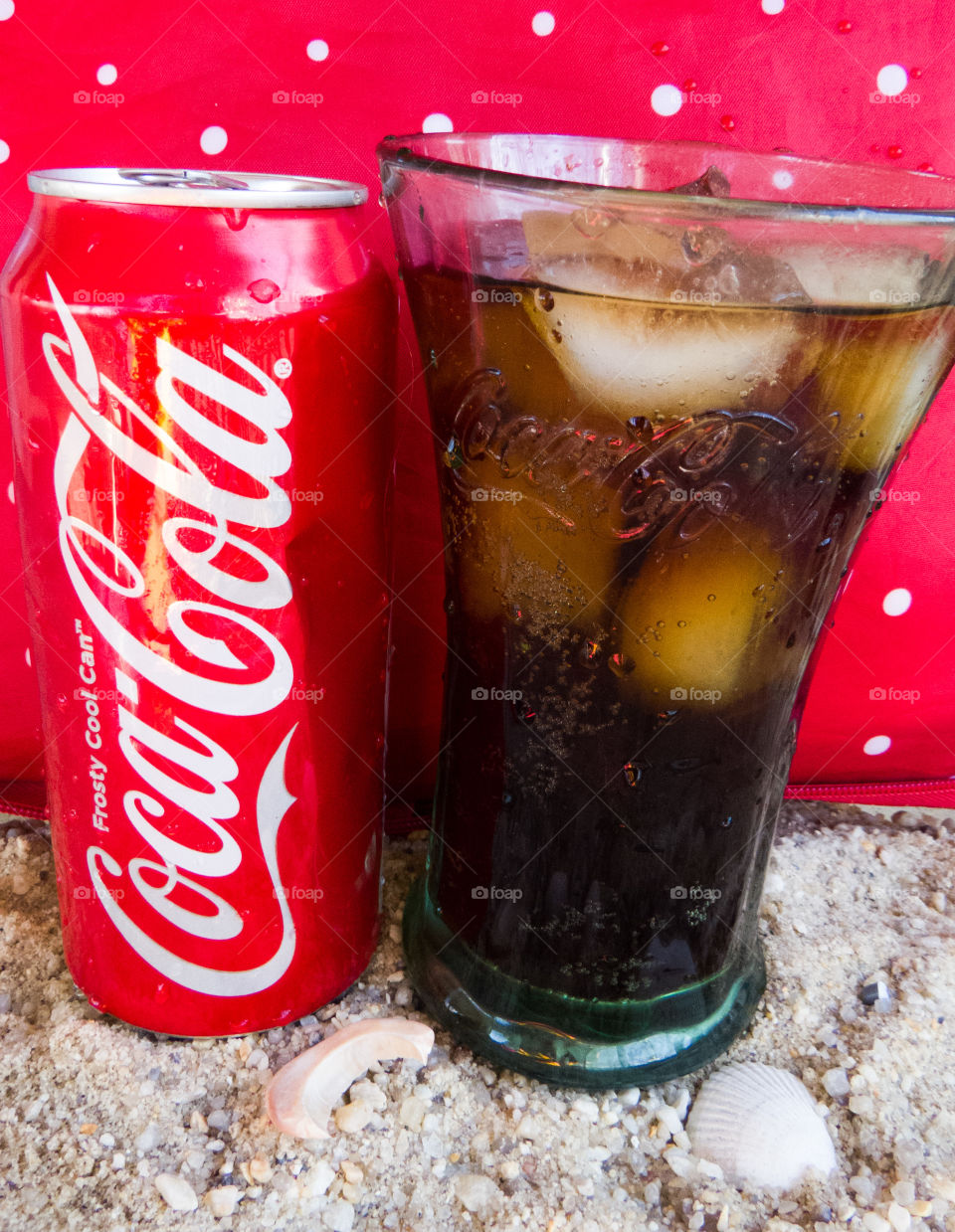 Coca-Cola can with full glass of coke in the sand with a red background.