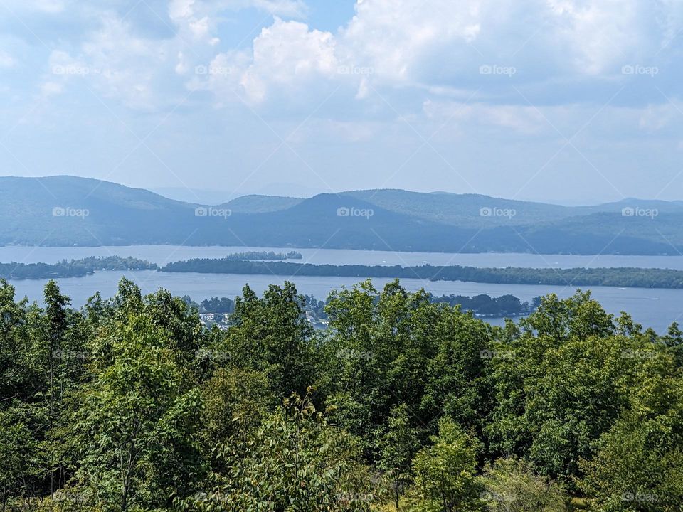View of Lake George from Buck Mountain in the Adirondacks. Trees in the foreground, the lake in the midground, and other mountains in the background. A cloudy sky.