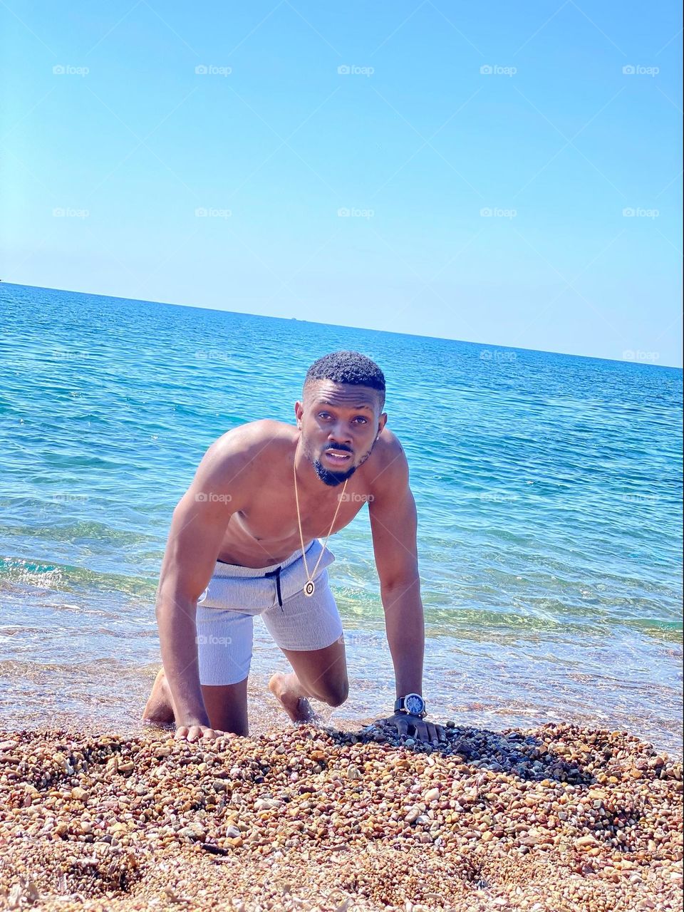 A muscular man with dark skin crawls across a sunlit beach, sweat glistening on his body. His intense gaze is fixed ahead, each deliberate movement showing determination, framed by the waves.