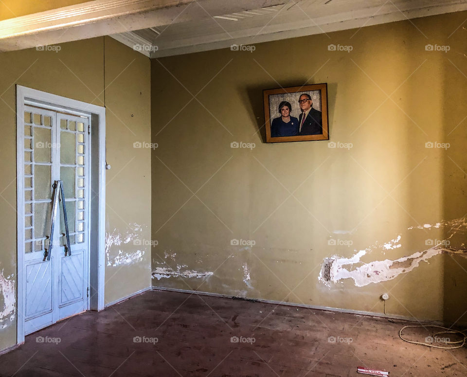 A photograph of a middle aged couple, is the only remaining item in an otherwise empty room. The walls damaged and the floor dirty. Leading out of the room there is a set of Art Deco style doors, crying out for restoration 