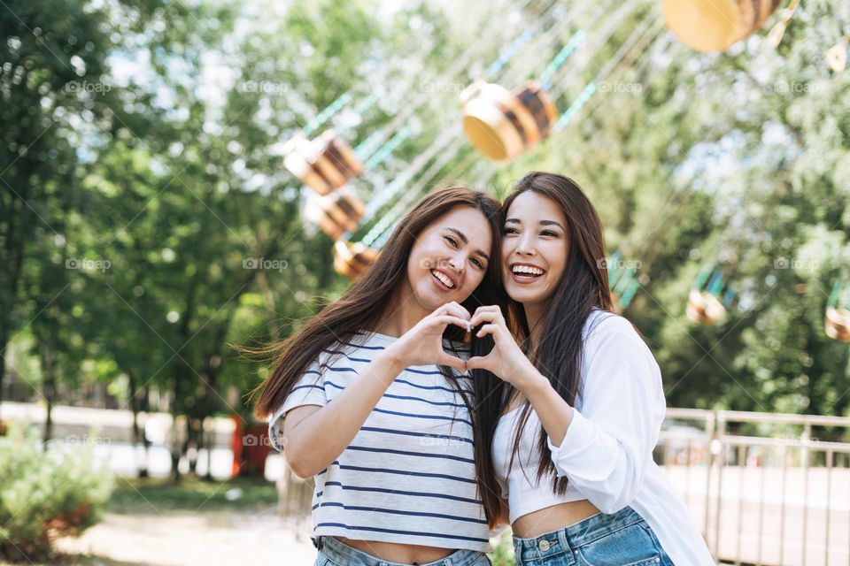 Young women with long hair friends having fun showing heart with their hands at amusement park