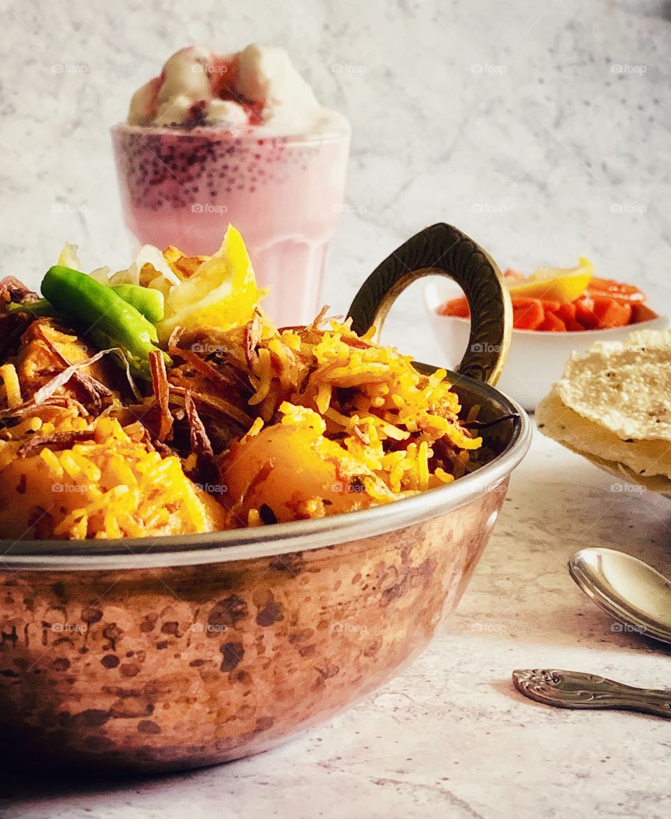 Chicken biryani in a copper pan served with rose milkshake served with condiments 