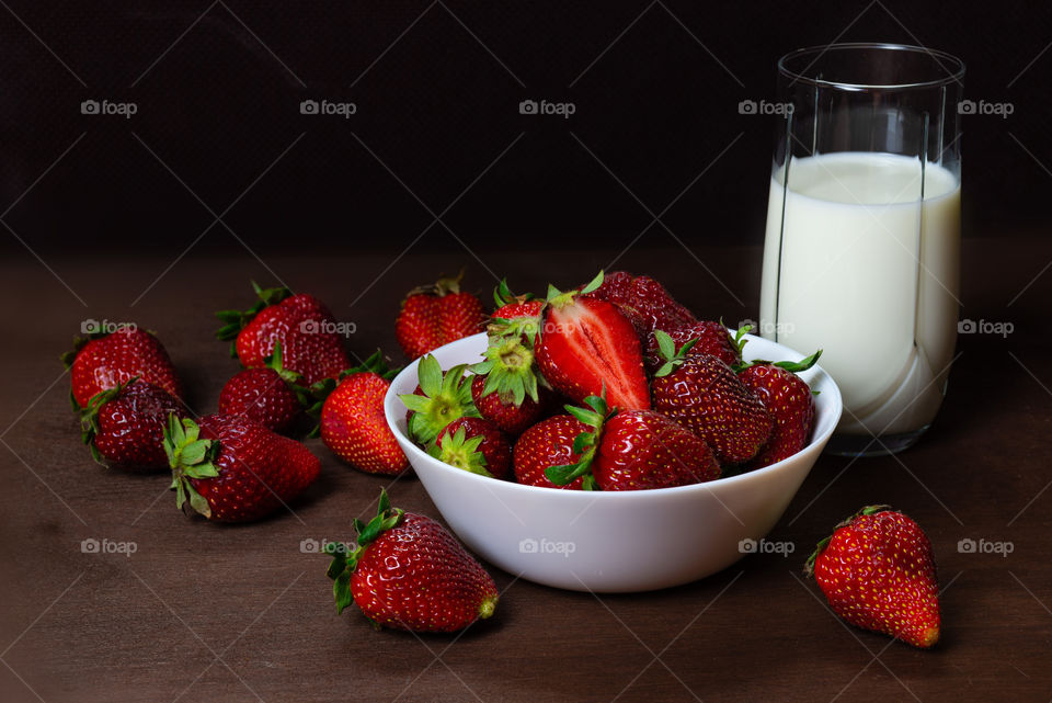 ftesh strawberries in ceramic bowl and a glass of milk on dark wooden background