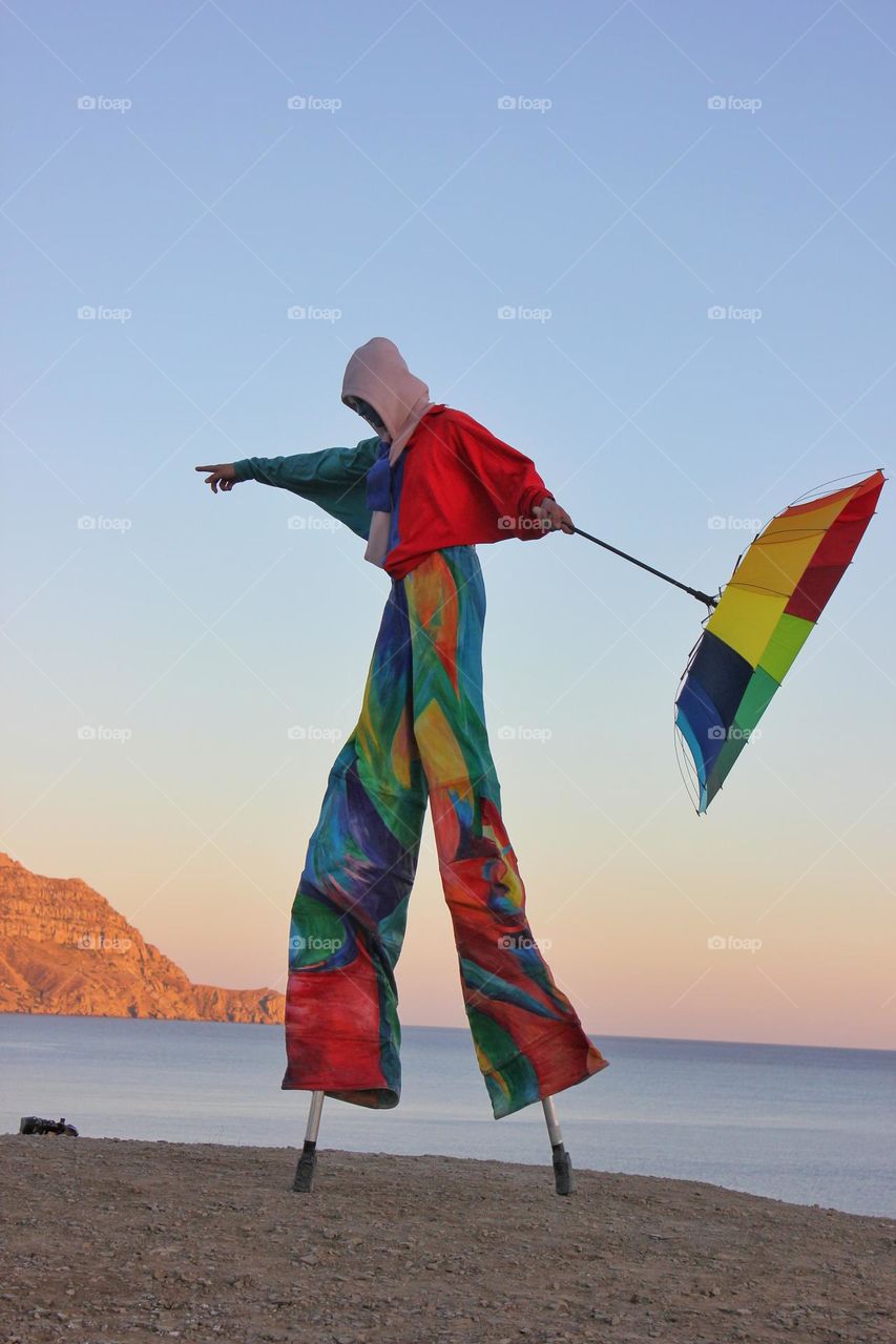 A man on stilts in colored clothes with an umbrella walks along the seashore in the evening. Clothing for the festival.