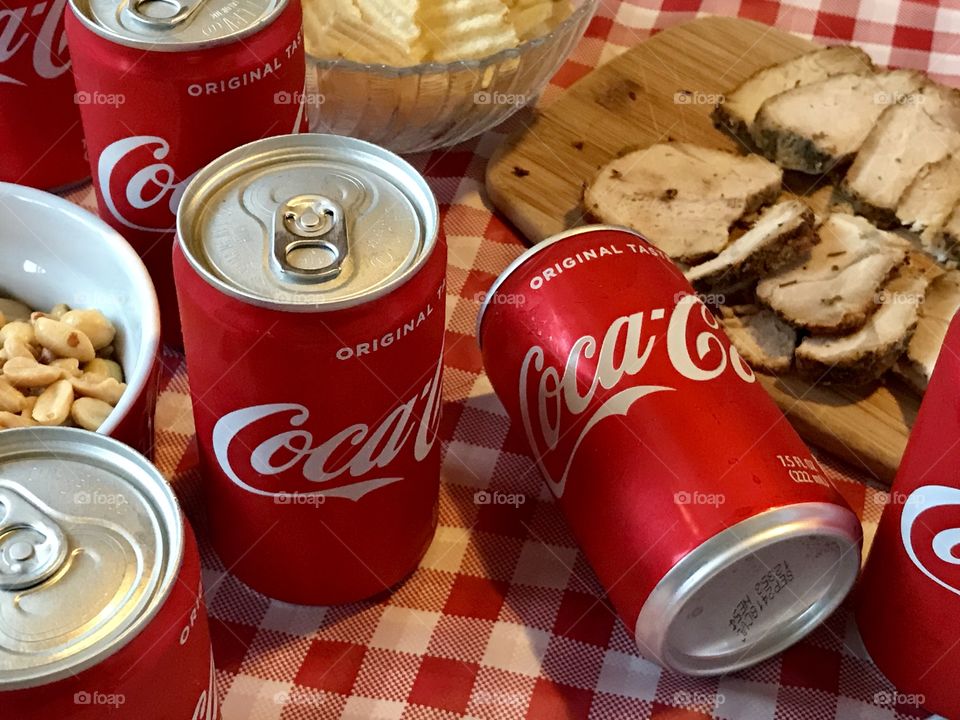 Cans of Coca-Cola on a red and white checkered background with barbecue food and potato chips 