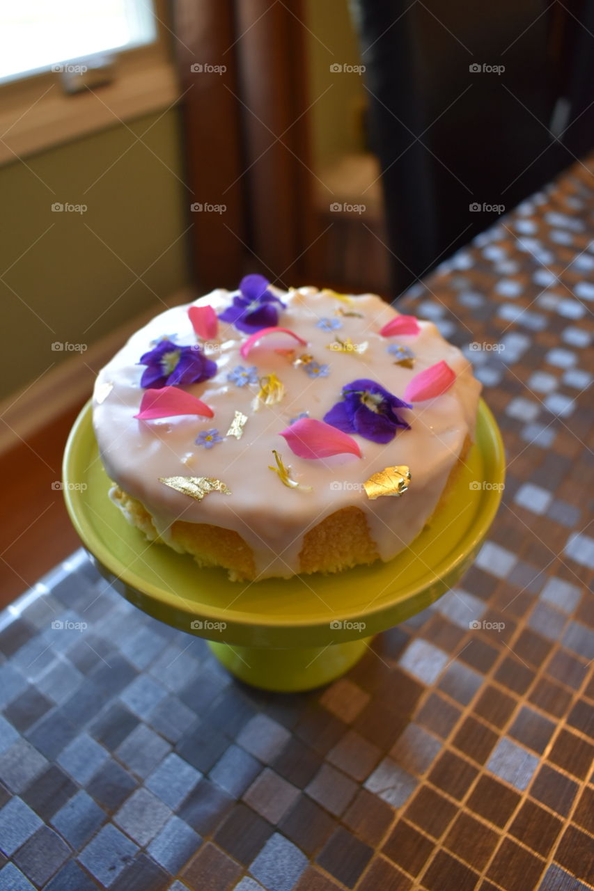 A small cake decorated with purple and pink flowers and gold leaf sits atop a yellow/ green cake stand. 