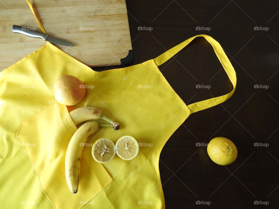 Yellow as a fall autumn season color with delicious vibrant and juicy lemons and bananas in a an apron pocket and grapefruit by a wooden cutting board and knife to cut the citrus fruits on a dining table.