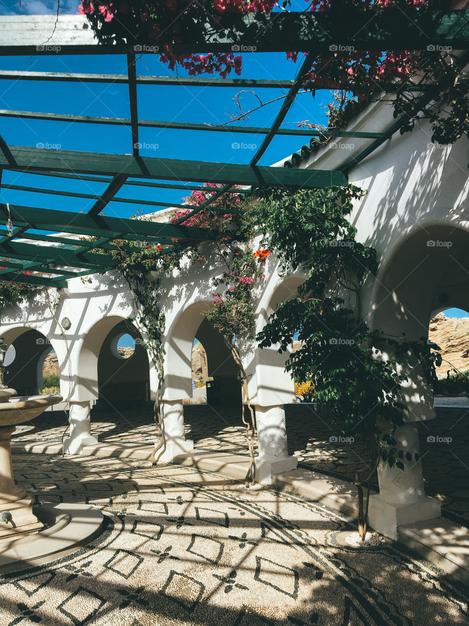 Shadows of leaves and wooden slats printed on the wall and on the floor , the walkway is beautifully designed with curved frames , blue sky . 