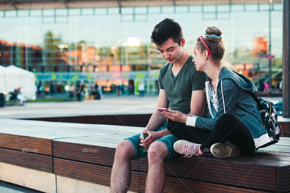 Couple of friends, teenage girl and boy, having fun together, using smartphones, sitting in center of town, spending time together. Real people, authentic situations